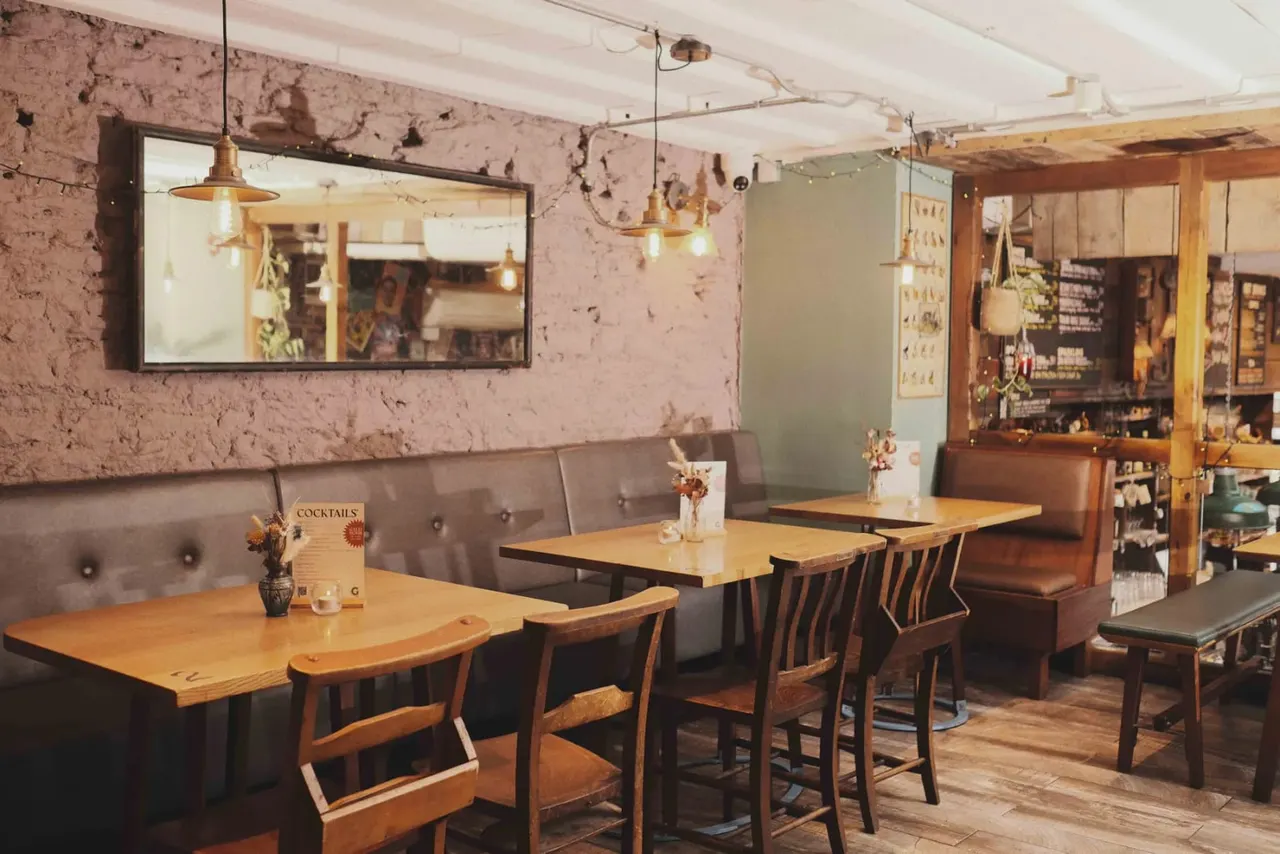 Industrial-style bar and restaurant interior with exposed pink brick walls, wooden tables and chairs, leather booth seating, and pendant lighting.