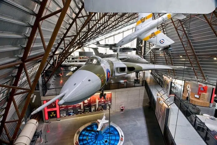 Military aircraft suspended from ceiling in large hangar museum space with exhibition displays below.