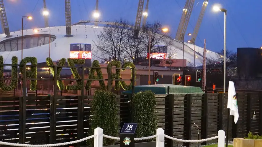 Twilight view of an adventure golf course with the Millennium Dome illuminated in the background and ivy-covered wooden fencing in the foreground.