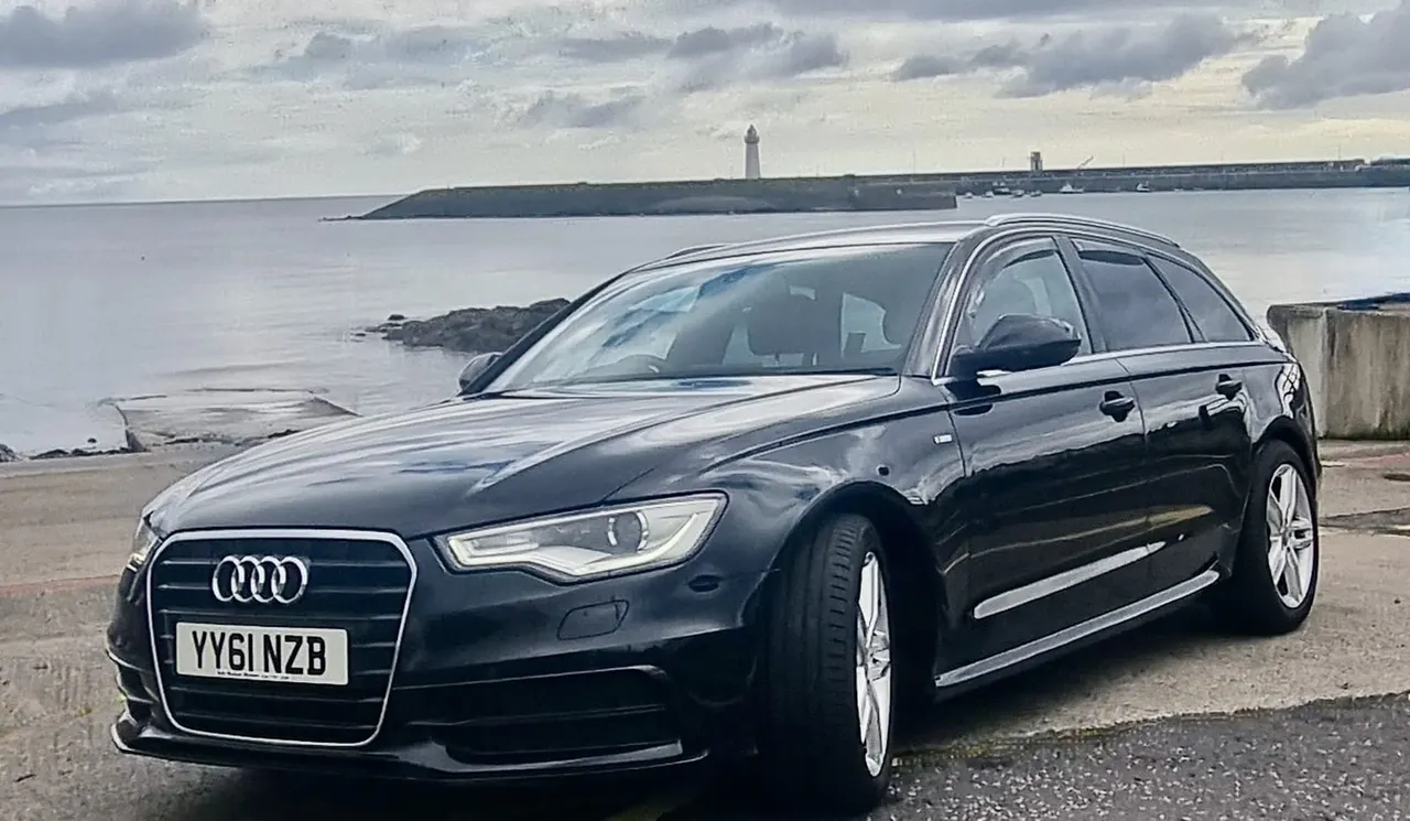 Dark Audi estate car parked on coastal road with calm sea, distant lighthouse and headland beyond.