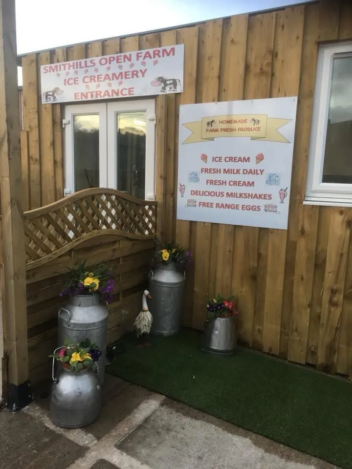 Entrance to a wooden farm building with signage for ice cream and farm produce, decorated with flowering planters and a wooden bench.