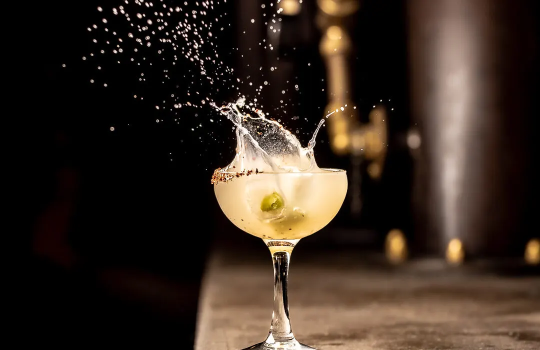A martini cocktail with olives splashing as liquid erupts from the glass against a dark background.