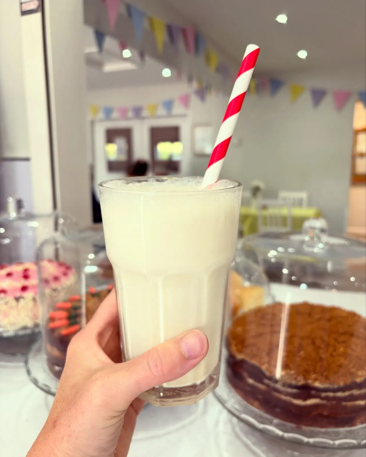 Hand holding a glass of milk with a red and white striped straw at a party table with cakes and bunting.