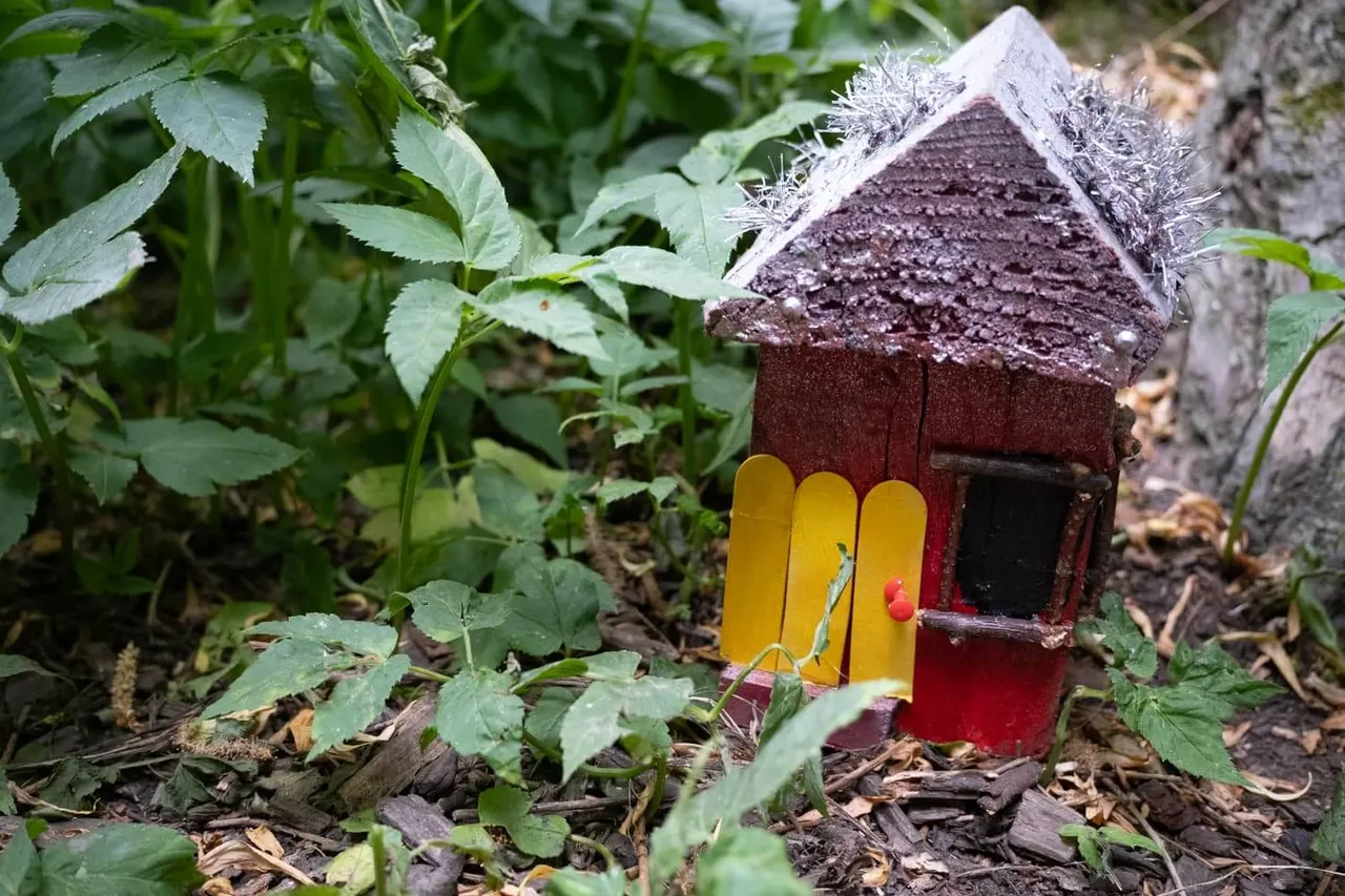 Miniature red and brown painted wooden house with yellow door nestled among green plants and tree bark in a woodland garden setting.