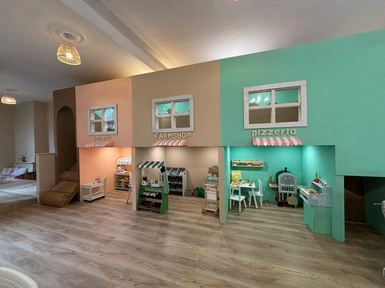 Indoor play area with three themed shop-front structures in pink, tan and turquoise, featuring a bakery, farmshop and pizzeria with miniature furnishings and accessories.
