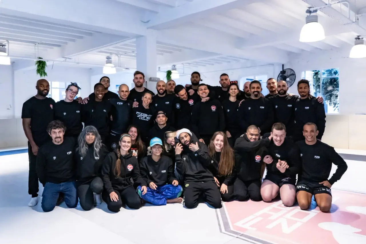 Large group of people in black branded clothing posing together on a padded gym floor in a bright, modern fitness studio.