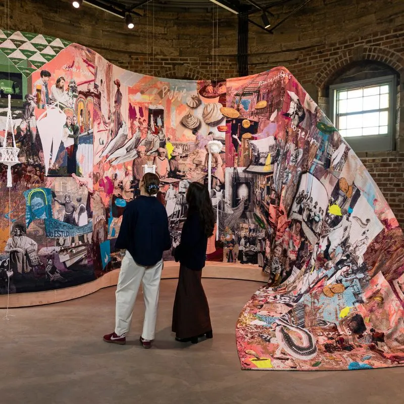 Two visitors viewing a large immersive art installation with collaged imagery in a brick-walled gallery space.