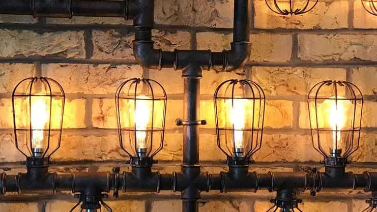 Industrial-style cage lights mounted on black pipework against a warm-lit stone brick wall.